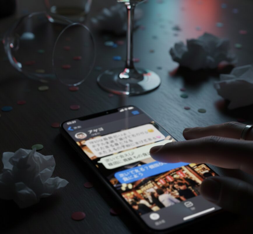 暗い室内で、散らかったテーブルの上に置かれたスマートフォンの画面。指が触れている画面には、メッセージアプリのやり取りが表示されているが、個人情報保護のため文字はぼかされている。背景にはひっくり返ったグラスと紙吹雪、ティッシュのくずが見える。