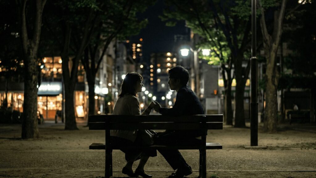 大阪市西区の靭公園（うつぼこうえん）周辺で、夜の闇に紛れてベンチで親密に手を握り合う男女のシルエット。周囲のおしゃれな店の明かりを背景に、浮気や密会デートの典型的な行動パターンを捉えた写真。