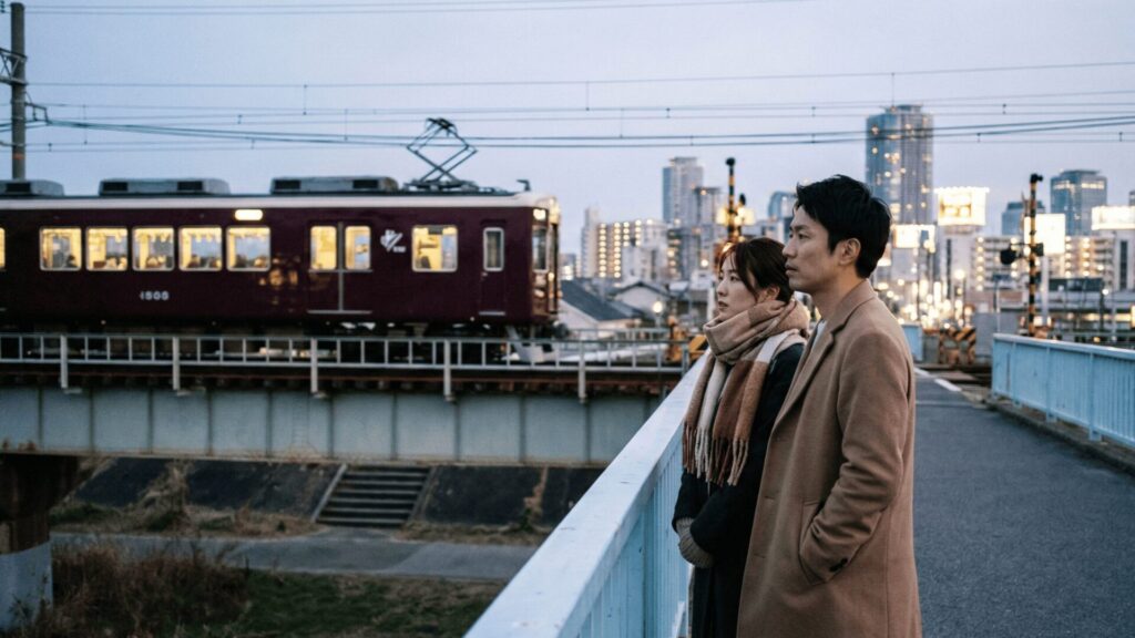 大阪府池田市の猪名川にかかる鉄橋近くで、通過する阪急電車と大阪方面の街明かりを見つめる日本人男女。年末年始の帰省時に、都市部へのアクセスの良さが浮気のきっかけになる地域特性を暗示している。