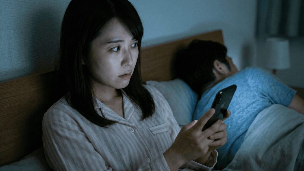 深夜の寝室で、隣で眠る夫に気づかれないよう、緊張した面持ちで夫のスマートフォンを手に取る女性。自分で浮気の証拠を掴もうとする「セルフ調査」の危険な一瞬。
