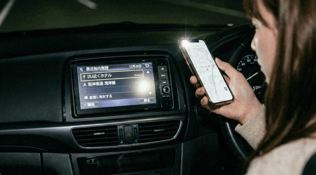 夜の車内で、女性がスマートフォンのライトをかざしてカーナビの目的地履歴を確認する様子。画面には「12月24日」の日付で「USJ近くホテル」の履歴がはっきりと残っており、大阪府堺市の男性がクリスマスに浮気をした動かぬ証拠を発見した瞬間。