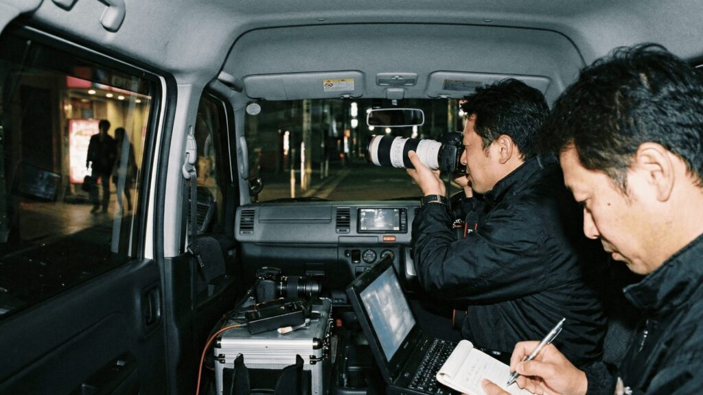 夜間の西宮市で、プロの探偵が車内から望遠カメラを使い、浮気の決定的な証拠を撮影している張り込み調査の様子