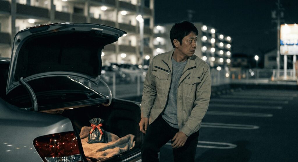 大阪府堺市の夜の駐車場で、浮気相手へのクリスマスプレゼントを車のトランクに隠し、周囲を警戒しながら振り返る作業着姿の男性。動かぬ浮気の証拠。