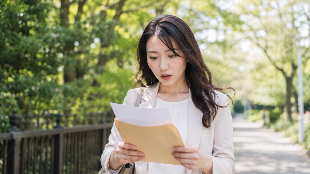 公園の遊歩道で書類を見ながら驚いた表情を見せる日本人女性。結婚前には知らなかった事実や婚前調査で発覚する情報を知った瞬間の心理を表現した写真風イメージ。